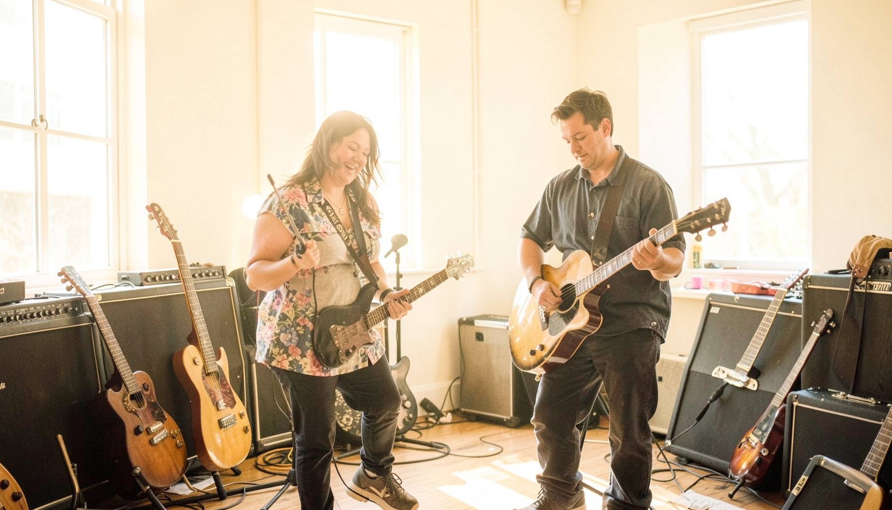 Two musicians in a sun-drenched rehearsal space with guitars leaning against amps, warm natural light streaming through large windows