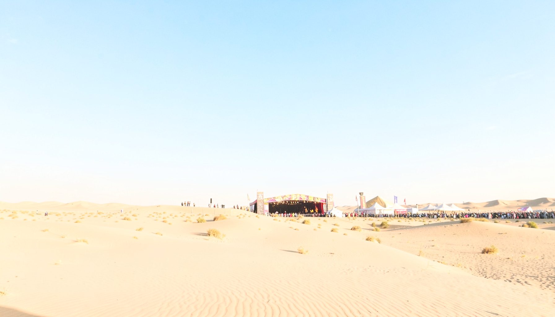 Bright desert landscape with a distant festival stage under a wide blue sky, golden hour light