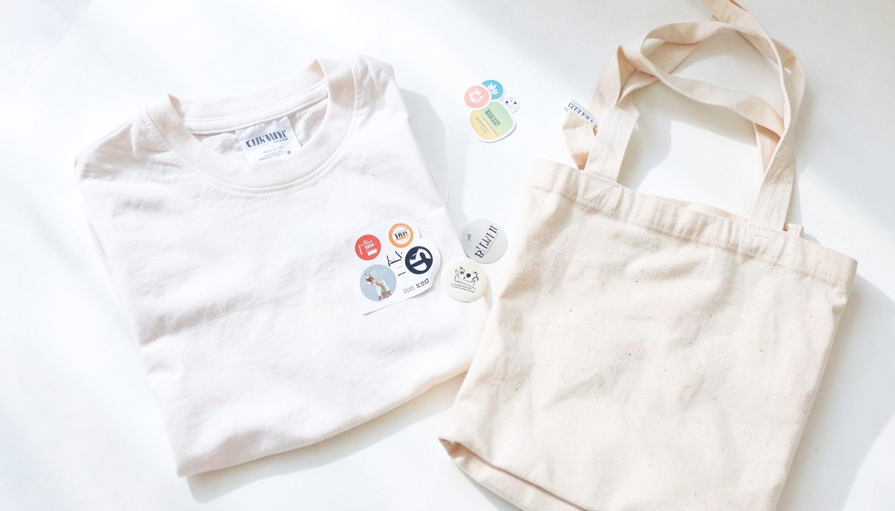 Bright, airy flat lay of band merchandise on a sunlit white surface: a folded tee, stickers, and a tote bag in soft neutrals
