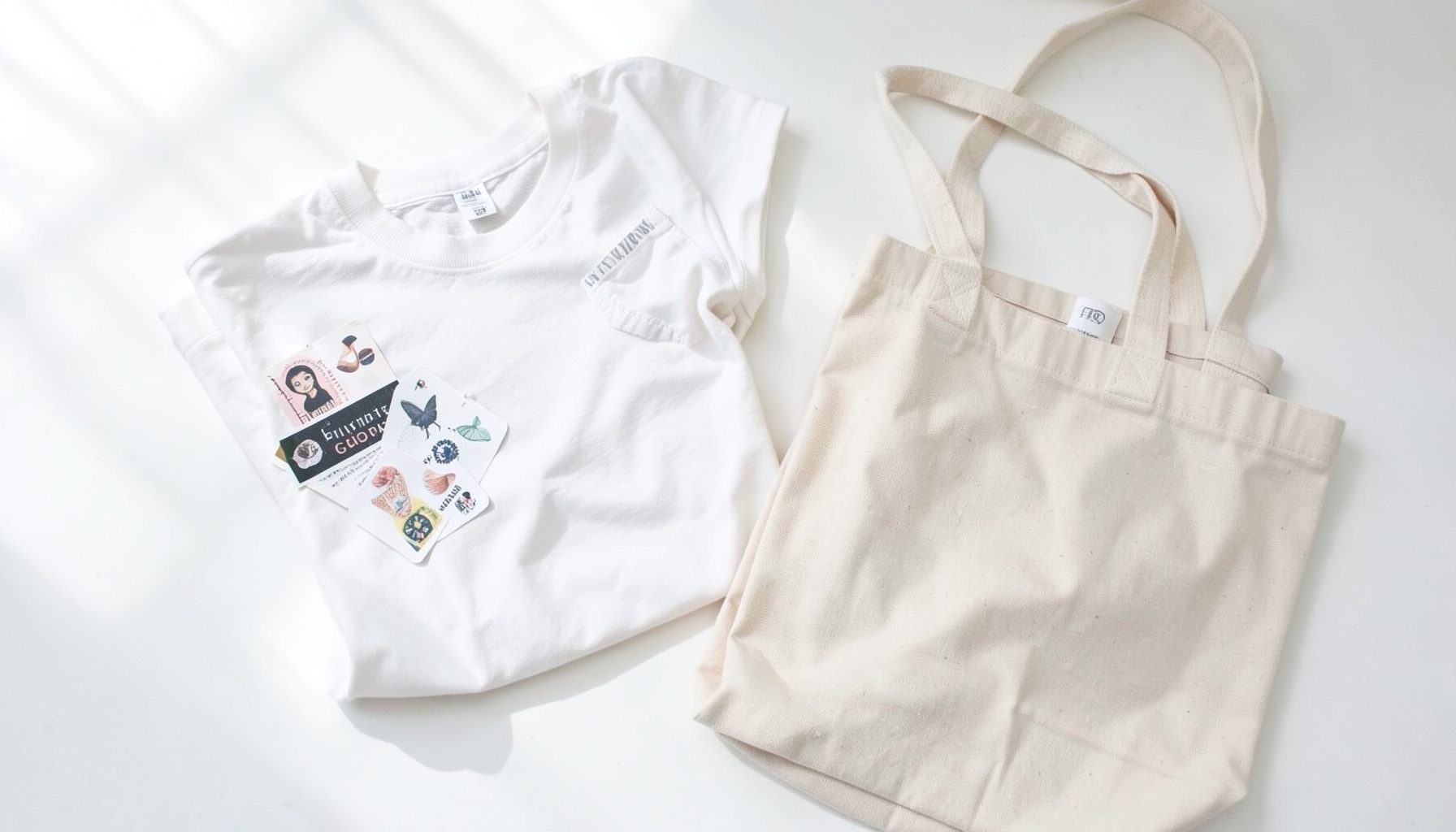 Bright flat lay of band merchandise on a sunlit white surface: a folded tee, stickers, and a tote bag in soft neutral tones