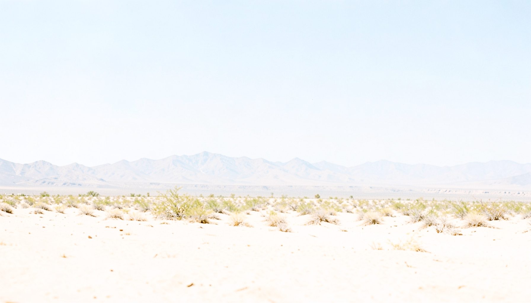 Bright, airy photograph of sunlit desert landscape with pale sand, distant low mountains, and a vast soft-blue sky, conveying warmth and open-road freedom